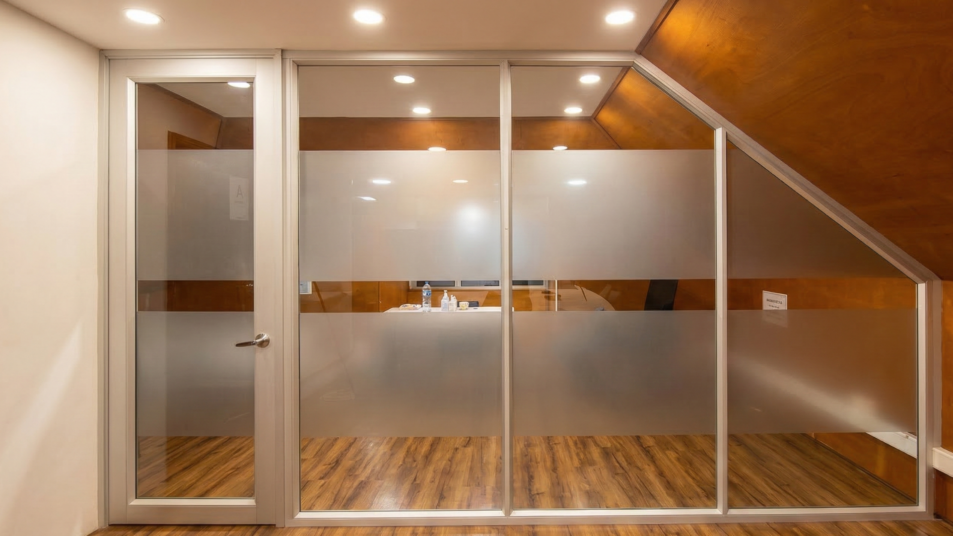 División en aluminio blanco con vidrio esmerilado y puerta abatible en oficina con techo inclinado en Colombia